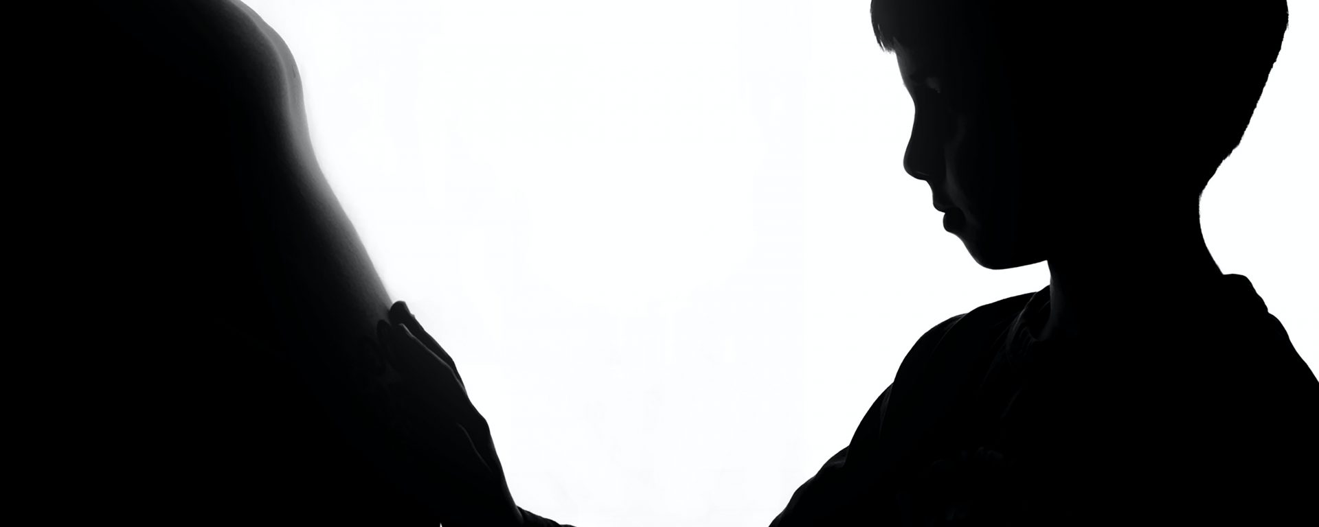 Boy with hand on pregnant womans stomach, both in silhouette.